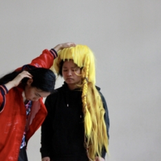 two female performer with asian backround standing in front of a light grey wall. one is wearing a bright red jacket and long brown hair. touching her ear with one hand, as the other hand rests on the other persons head. she is looking to the floor. the other person wears a massive yellow wig. the hair is structured in a long messy style falling over her left shoulder. she waers a black antifa-like hoddy and has her eyes closed.