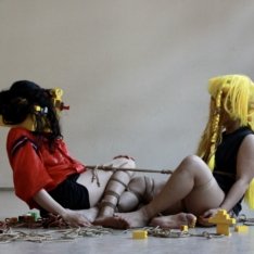 Two performers sitting close to each other in front of a cream color backround. their bare legs are tied together with a brown rope. the one on the left wears a bright yellow jacket. on her head a square cube build of yellow lego duplo stones. mixed with her back long hair. the other perfomer on the right a black training shirt, her light brwon skin of her arms visible, and her head covered with a massive bright yellow long wig. their chests are connected with a thick rope into which they are leaning backwards holding each other with their weigt.
