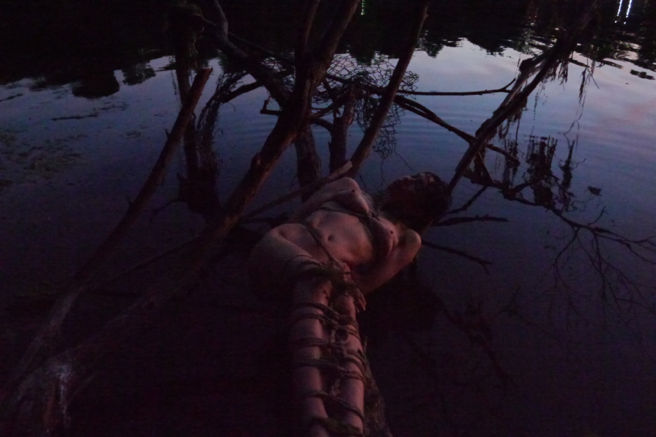 female lying tied in ropes on tree trunk in water its after sunset and there theere are more shadows than light